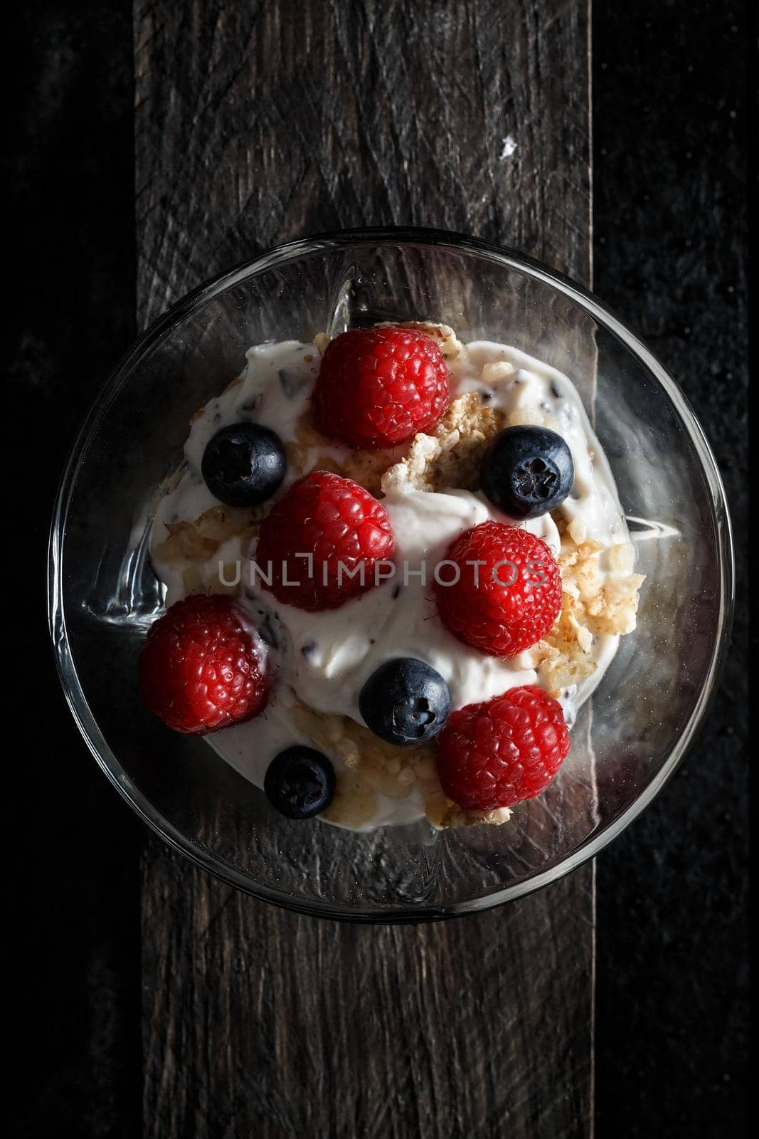 Raspberries, blueberries, cereals and yogurt in a glass bowl on old wooden boards. Healthy breakfast for a healthy life. Vertical image view from above.