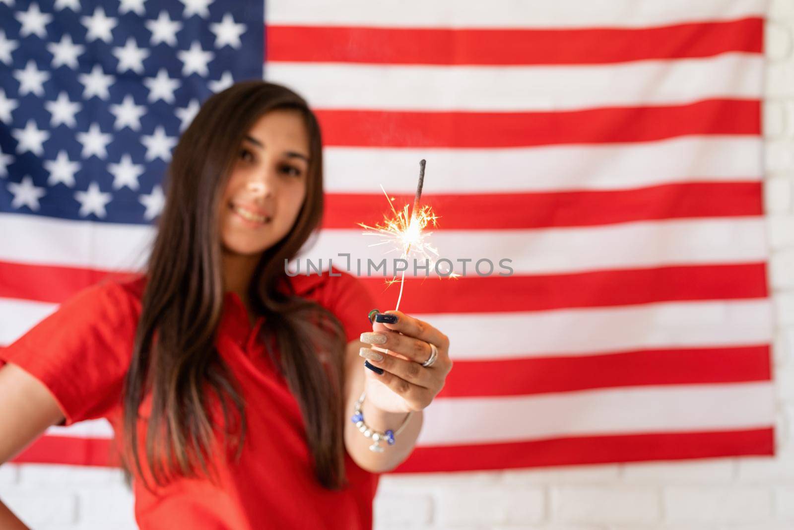 Independence day of the USA. Happy July 4th. Sparkling Bengal fire in caucasian woman hand. Beautiful woman holding a sparkler on the US flag background