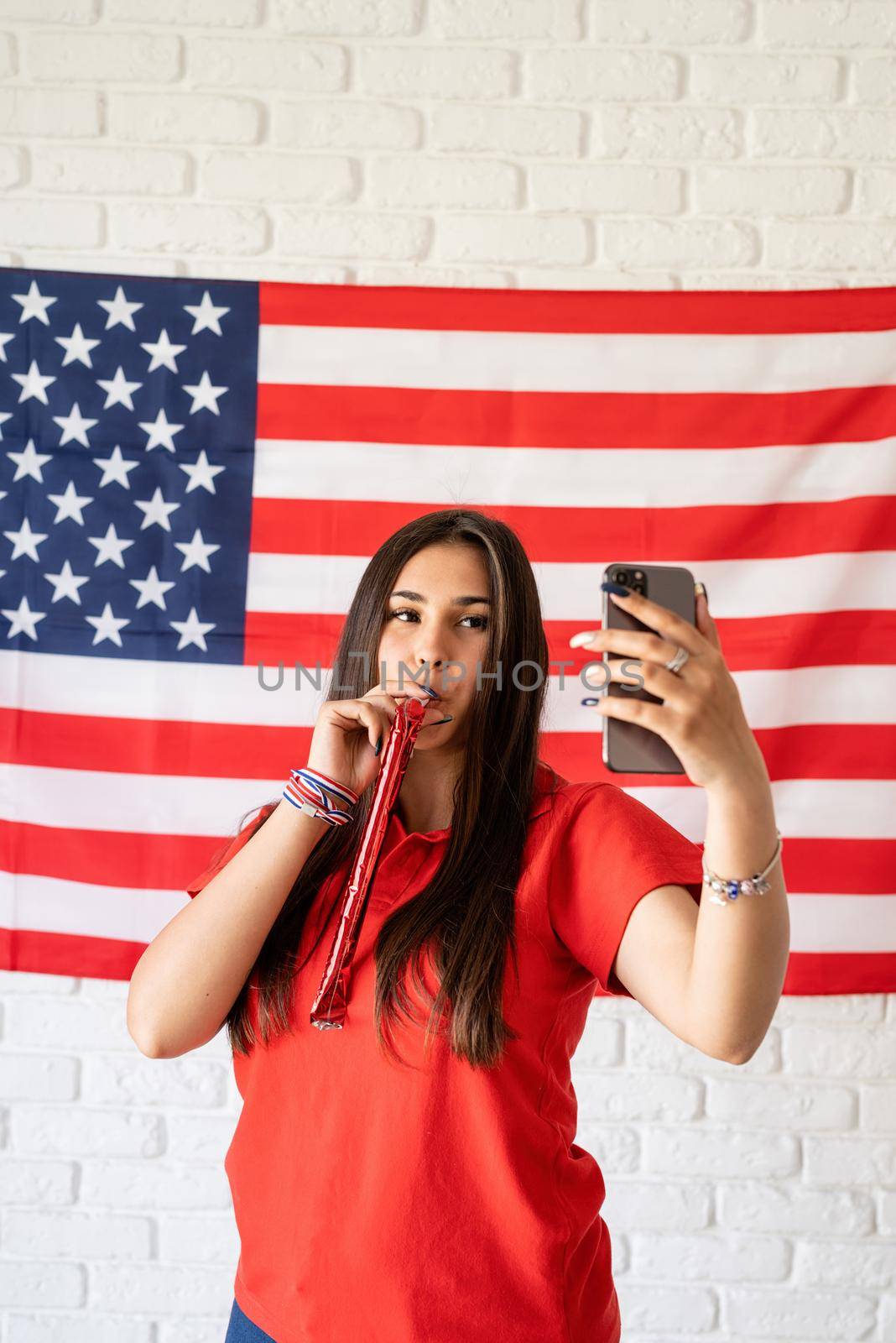 Independence day of the USA. Happy July 4th. Beautiful woman with a noisemaker taking a selfie on the USA flag background