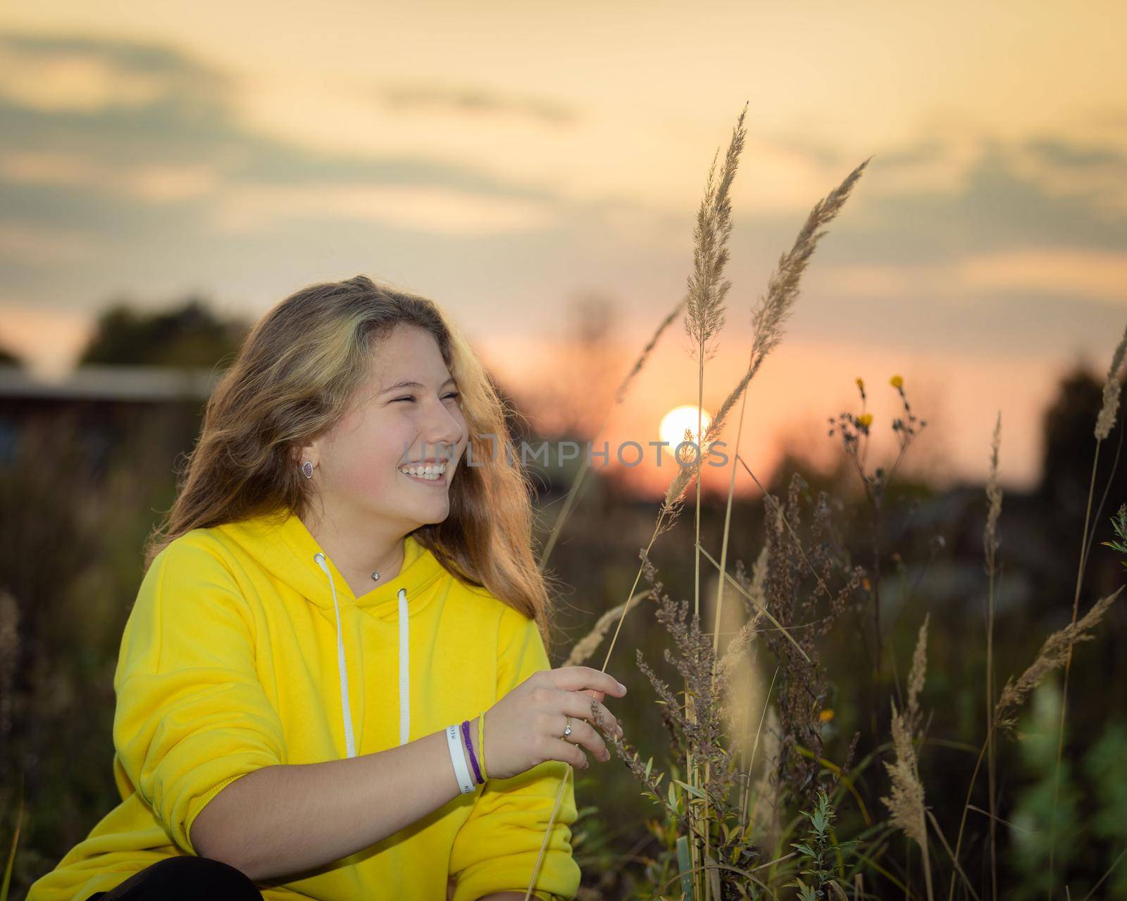 A young girl with long hair on a meadow with tall grass in the rays of the setting sun. Blonde hair. Yellow jacket.