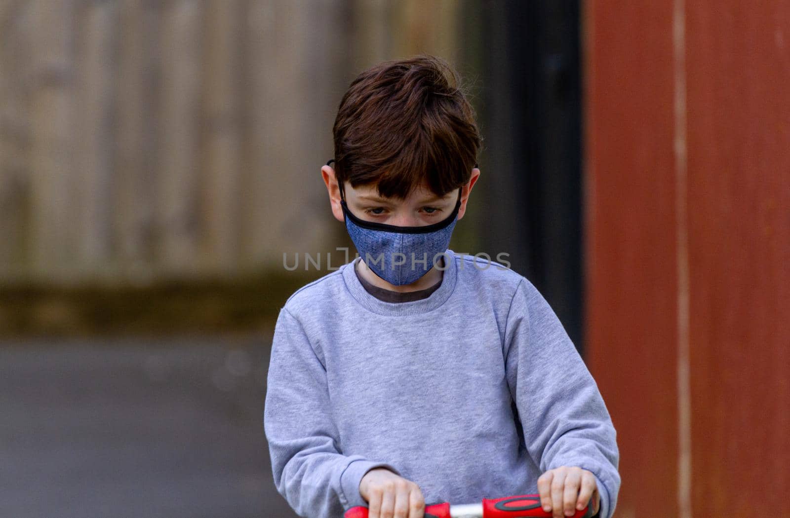 Portrait of a redhead boy wearing a face mask, riding a red scooter on a tar path surrounded by wood fences