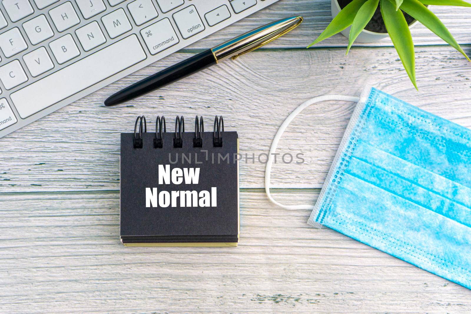 NEW NORMAL text with notepad, safety face mask, keyboard, fountain pen and decorative plant on wooden background. Covid 19 and coronavirus concept