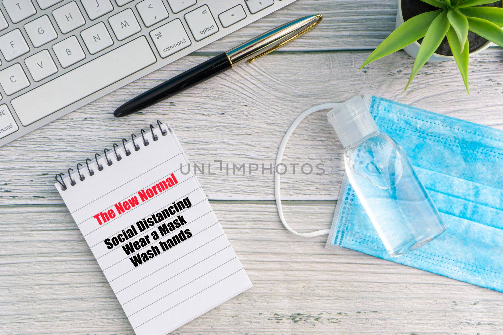 THE NEW NORMAL text with notebook, safety face mask, keyboard, fountain pen, hand sanitizer and decorative plant on wooden background. Covid 19 and coronavirus concept