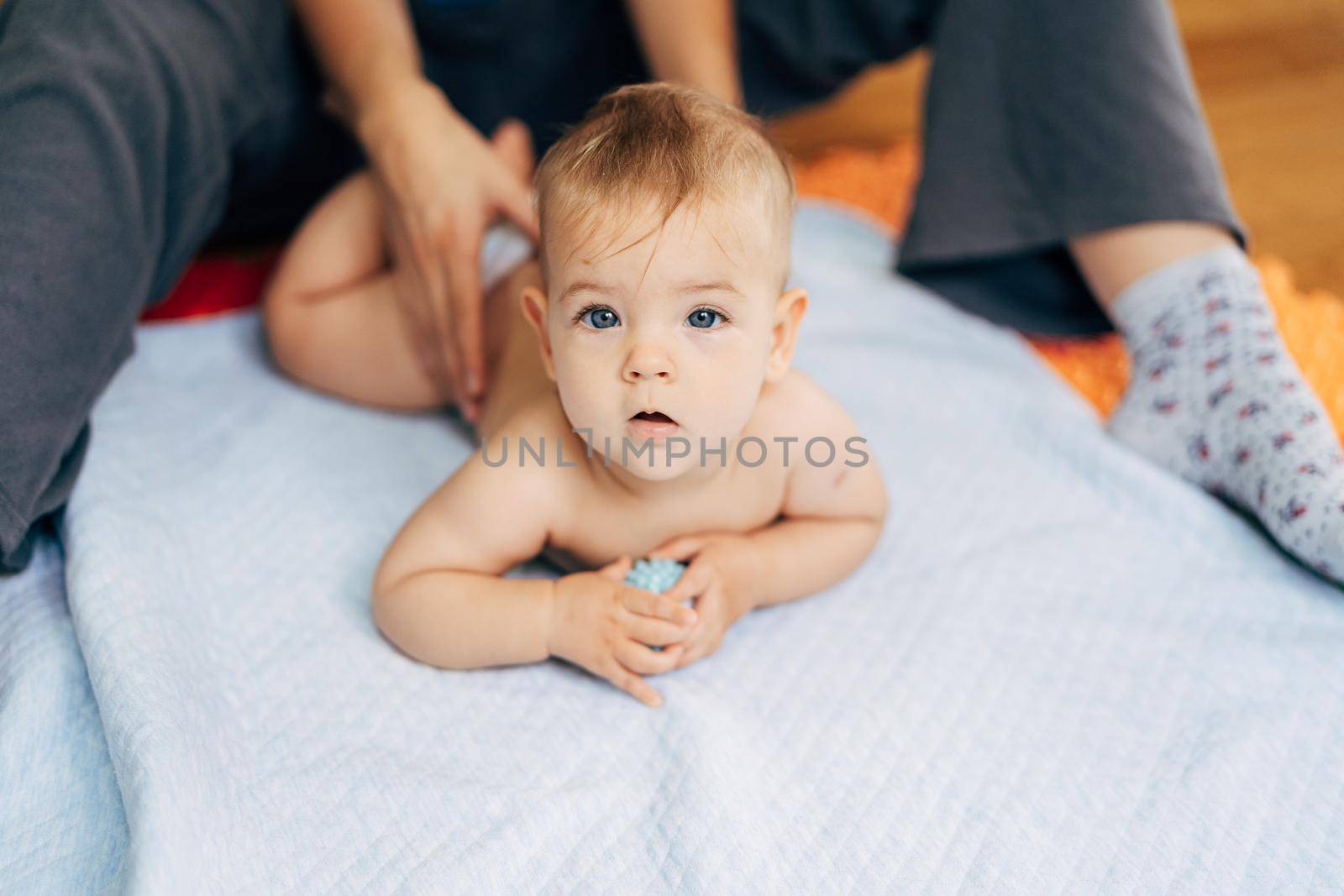 Tiny baby in diapers lies with his tummy on a blue blanket holding a toy in his hands. Mom's hands hold the baby by the back. High quality photo