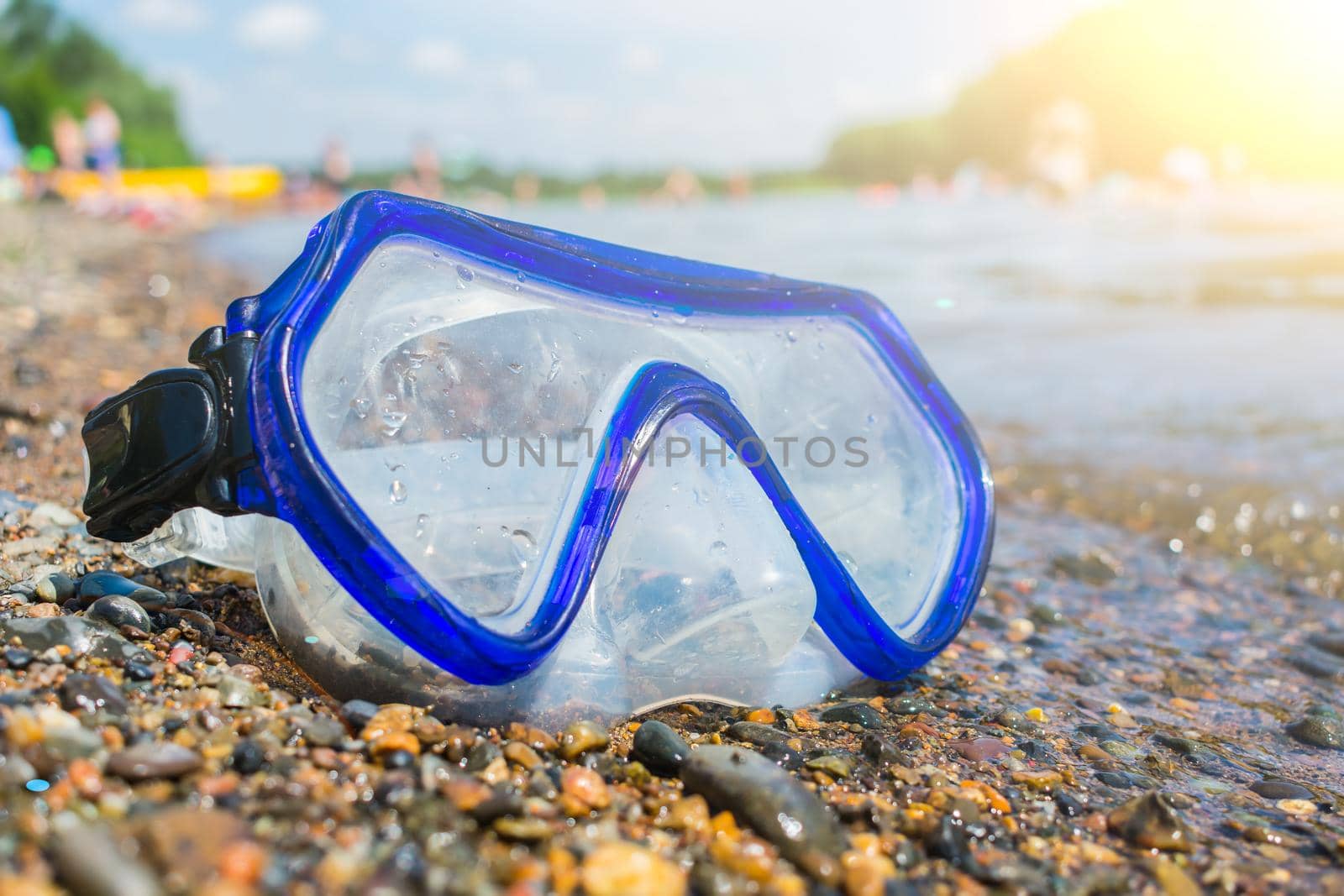 A swimming mask lies on a public beach near the water with people resting in the background