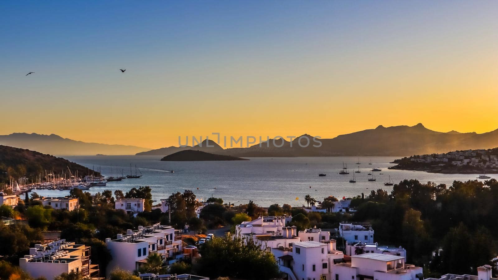 Bright colorful sunset in the beautiful bay of the Aegean sea with islands, mountains, boats and birds in the sky . Summer holiday concept and travel background