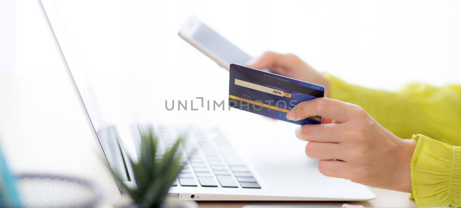 Closeup young asian business woman using smart phone and holding credit card while online shopping and payment with laptop computer on desk at home, female holding debit card, communication concept.