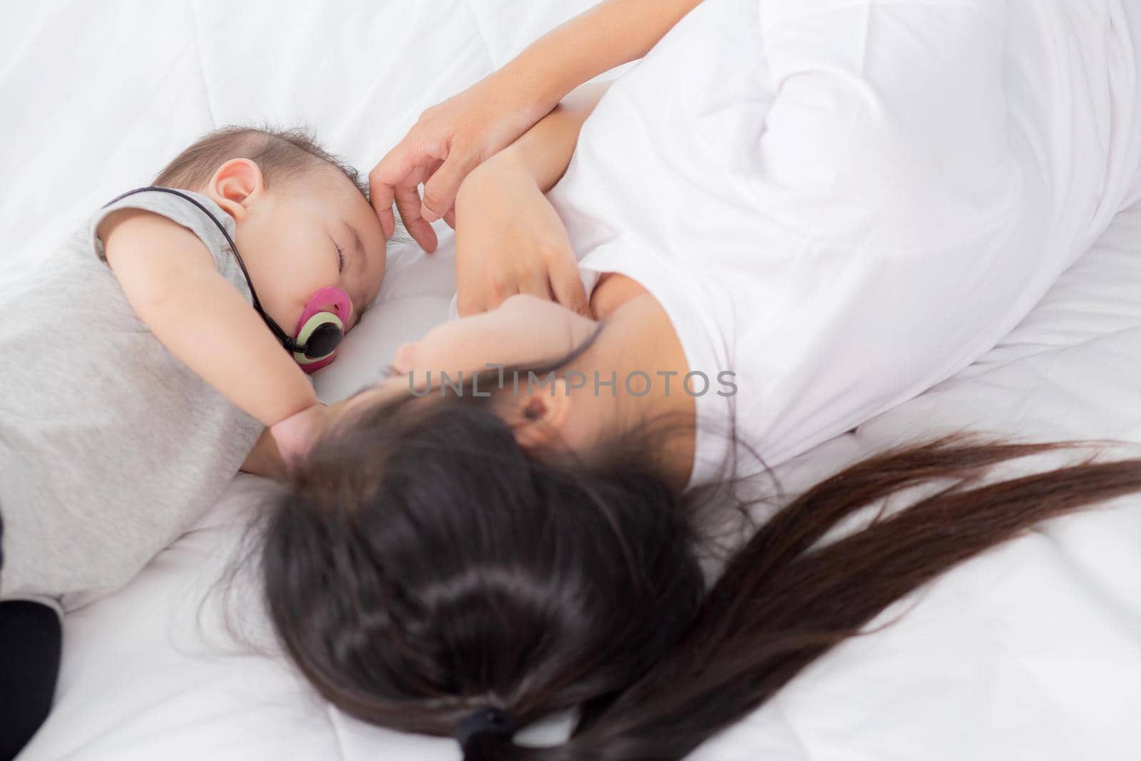 Young asian mother touch head of little baby girl with tender on bed in the bedroom, mom love newborn and care, woman with expression with child together, parent and daughter, family concept.