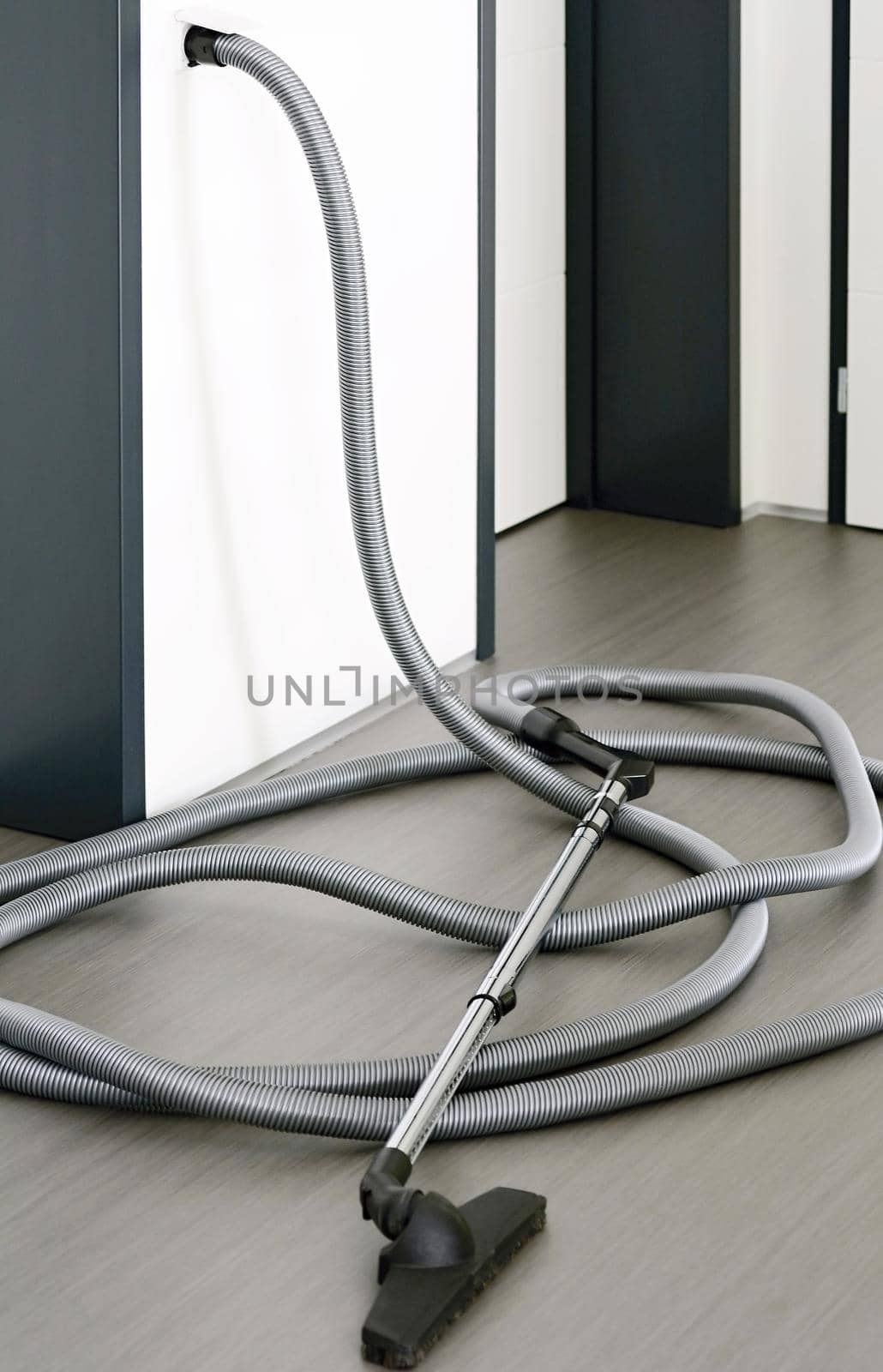 Long central vacuum cleaner hose laid on the grey floor in the room. Hose of central vacuum cleaner plugged into a wall inlet valve socket.