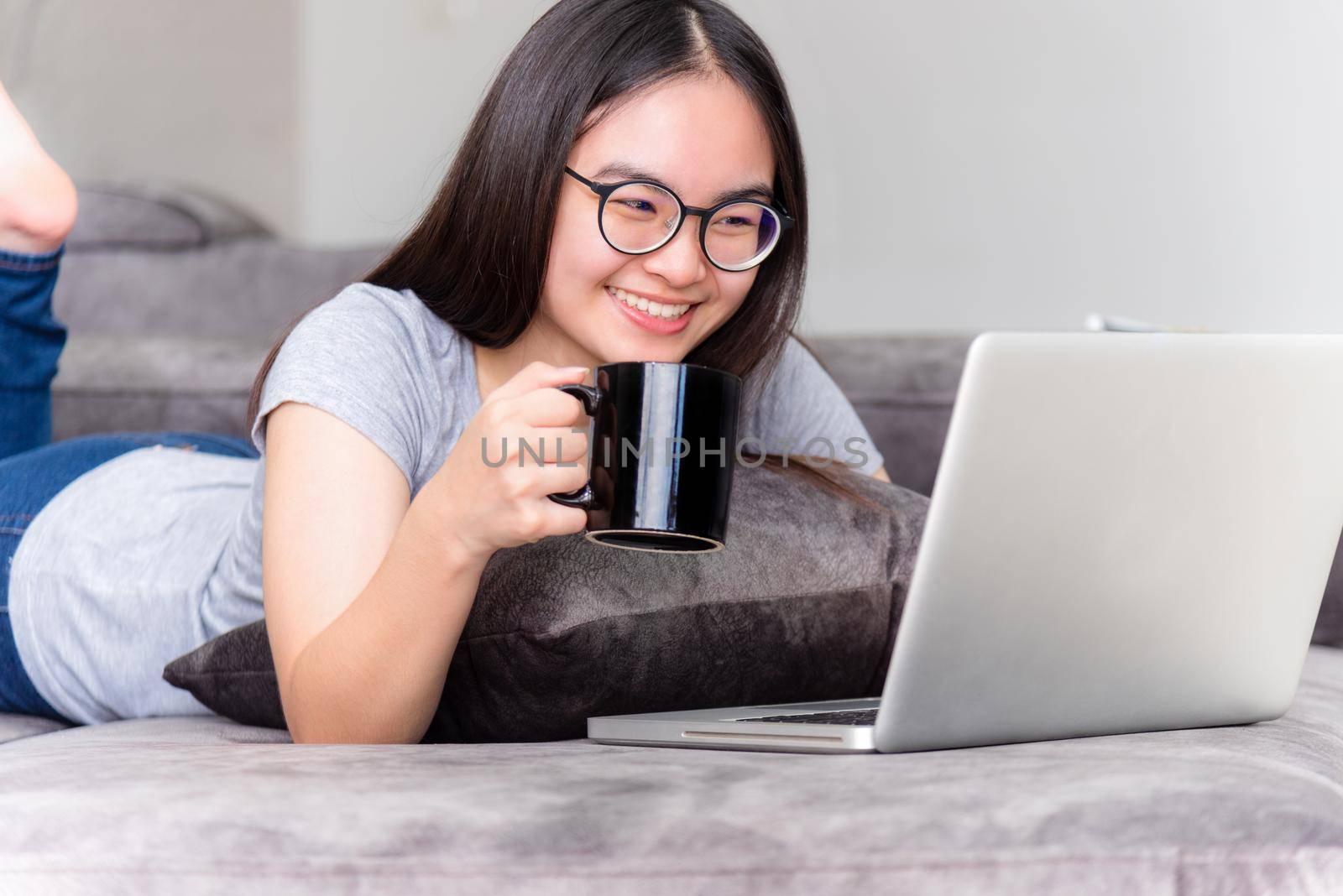Portrait beautiful Asian young woman lying relax hold coffee cup work with a laptop, Cute girl teens looking at monitor smile happily on the sofa working remotely with an internet computer from home
