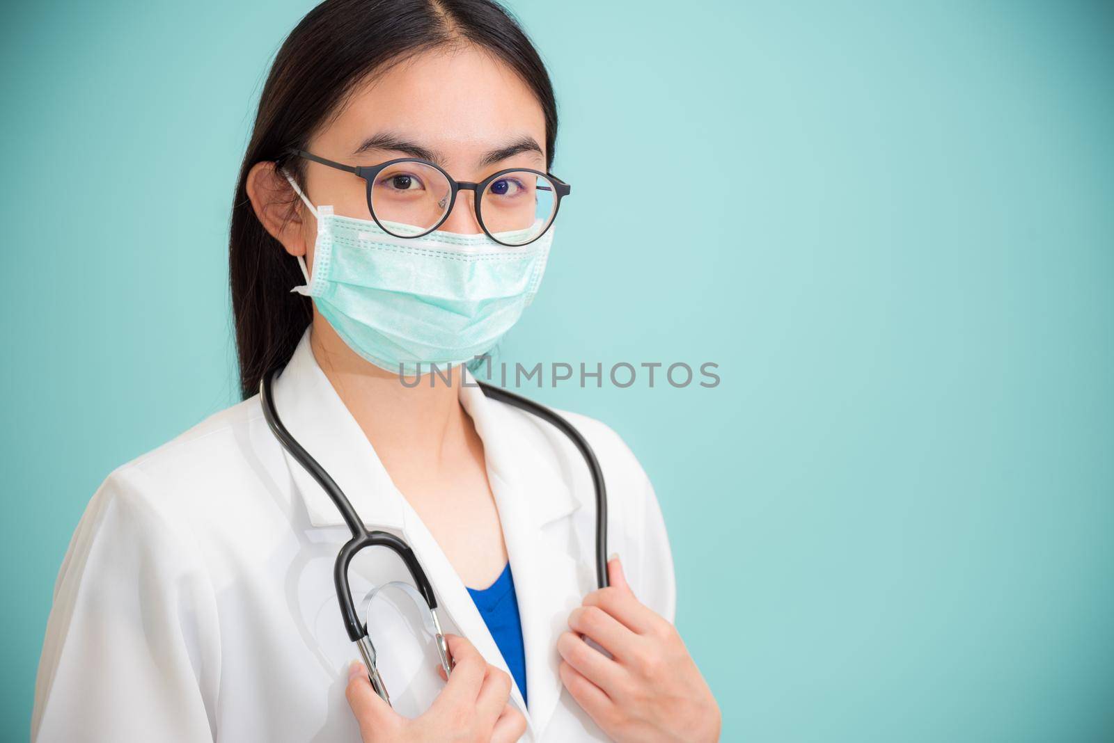 Studio portrait beautiful Asian young woman doctor with stethoscope in white uniform wear glasses and green mask to protect Corona Virus for health looking at camera on green copy space background