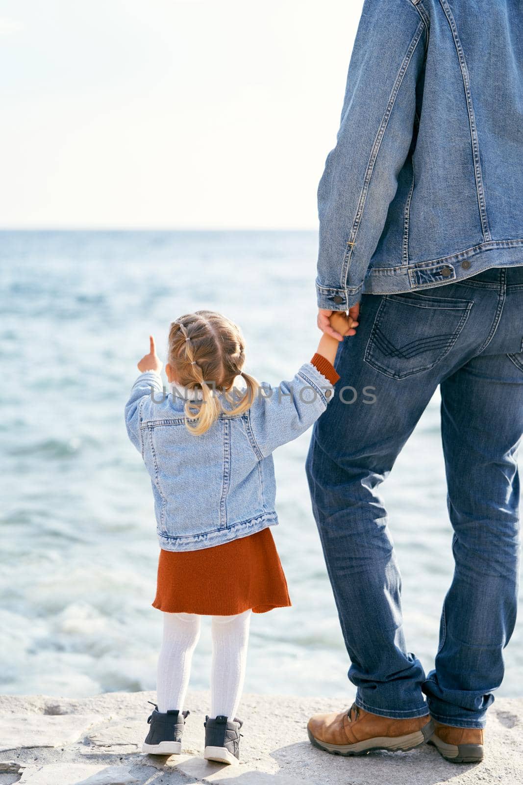 Dad in a denim jacket and jeans holds the hand of a little girl who points to the sea, standing on the stones by the water. Back view. High quality photo