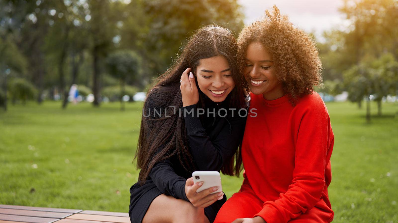 Diversity and feminity concept. Happy smiling Confident young mixed race women sitting bench park outdoor Asian and african american female resting on nature summer day using smartphone.