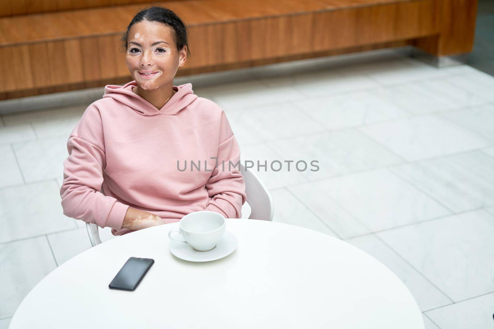 Black african american woman with vitiligo pigmentation skin problem indoor dressed pink hoodie sitting table high angle view