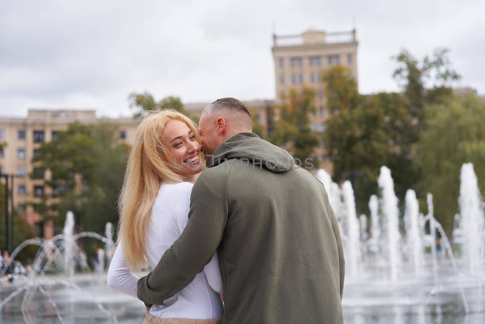 Romantic walk. Young couple in love embracing near fountain in urban park, man whispering compliments to his beautiful girlfriend