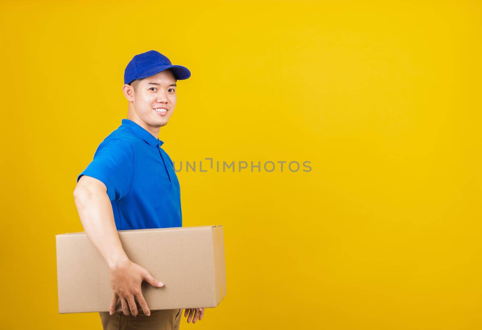Portrait excited attractive delivery happy man logistic standing smile wearing blue t-shirt and cap uniform holding parcel box looking to camera, studio shot isolated on yellow background, side view