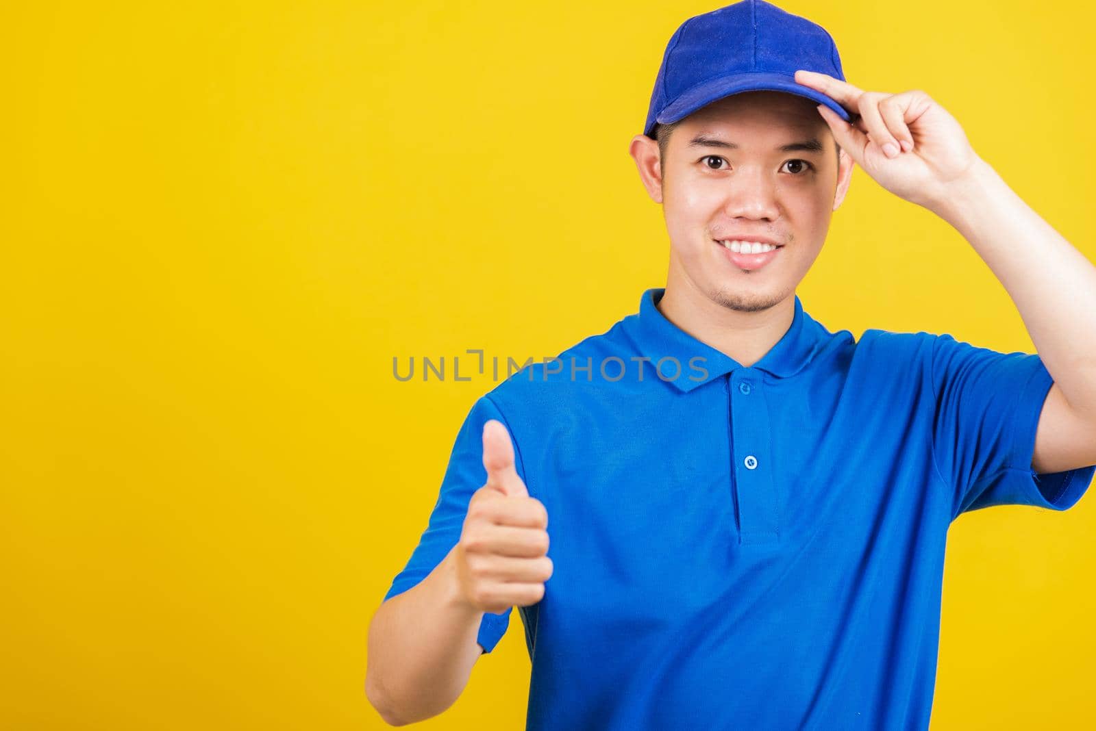 Portrait professional attractive delivery happy man standing he smile wearing blue t-shirt and cap uniform showing thumb up gesture looking to camera, studio shot isolated on yellow background