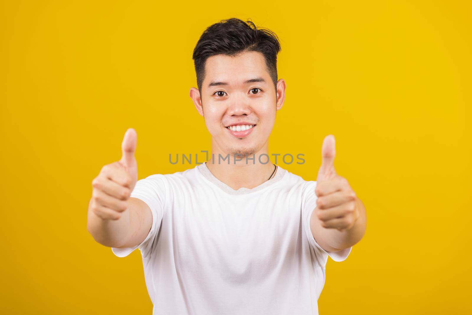 Asian handsome young man smiling positive showing thumbs up gesture good or like sign looking to camera, studio shot isolated on yellow background