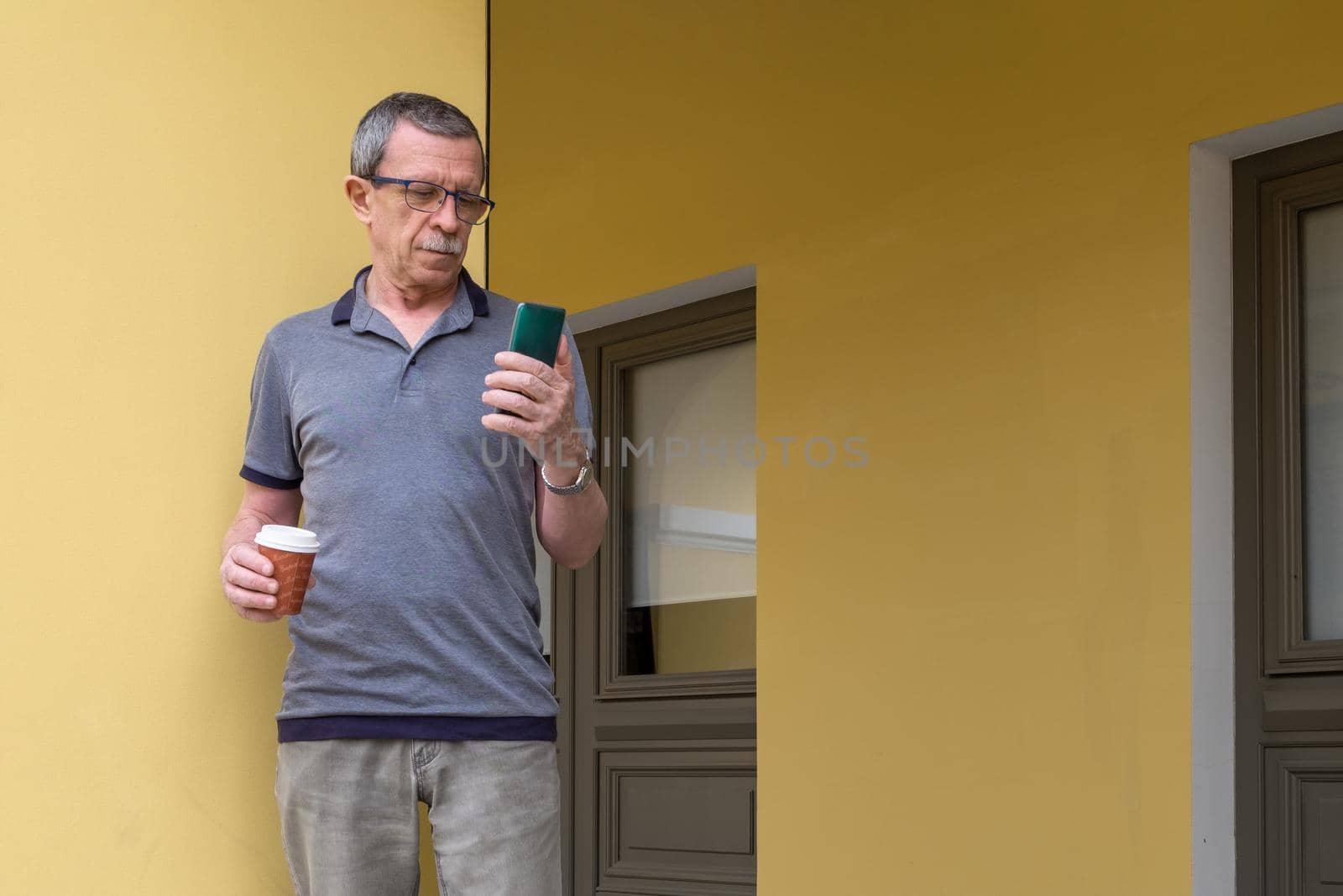An adult retired man in gray T-shirt with glasses, mustache and wrinkles on his face stands against the background of a yellow wall and holds a smartphone and a glass of coffee to go. Selective focus