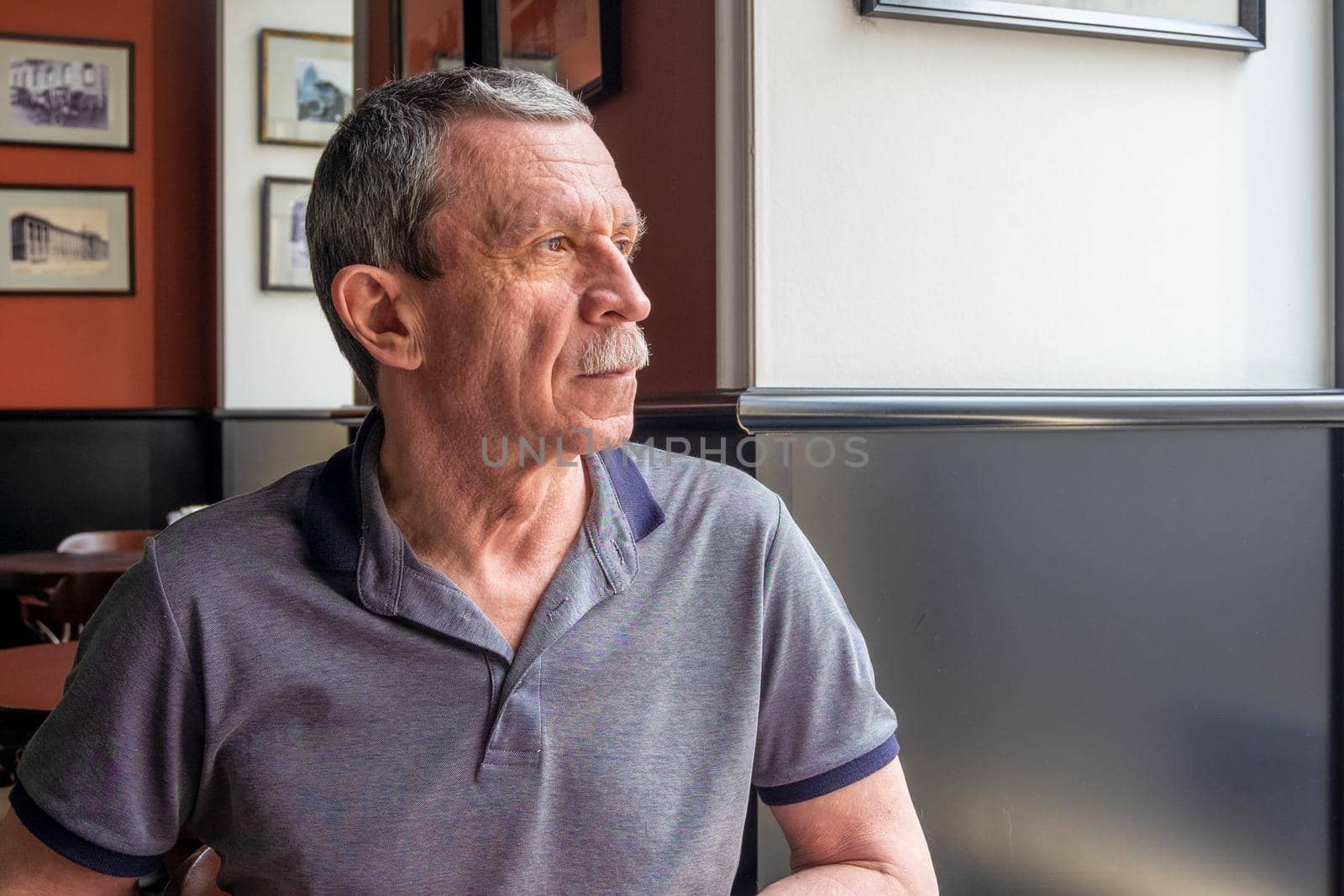 An adult Caucasian senior man in a gray T-shirt with a mustache and wrinkles on his face sits at a table in a cafe and looks out the window. Profile view. Selective focus.