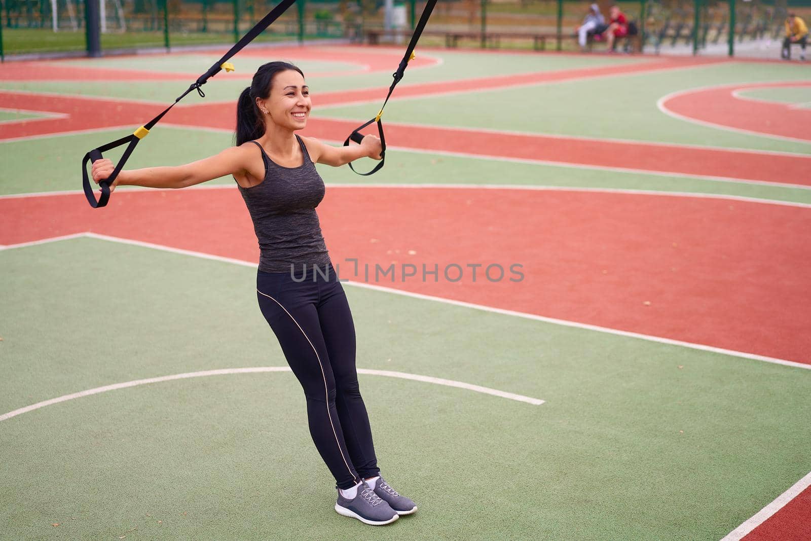 Girl athlete training using on sport ground. Mixed race young adult woman do workout with suspension system. Healthy lifestyle. Stretching outdoors playground. by andreonegin