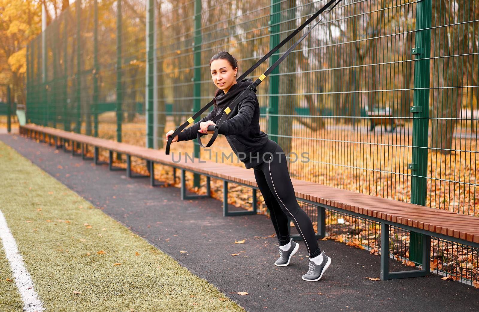 Girl athlete training using on sport ground. Mixed race young adult woman do workout with suspension system. Healthy lifestyle. Stretching outdoors playground. by andreonegin