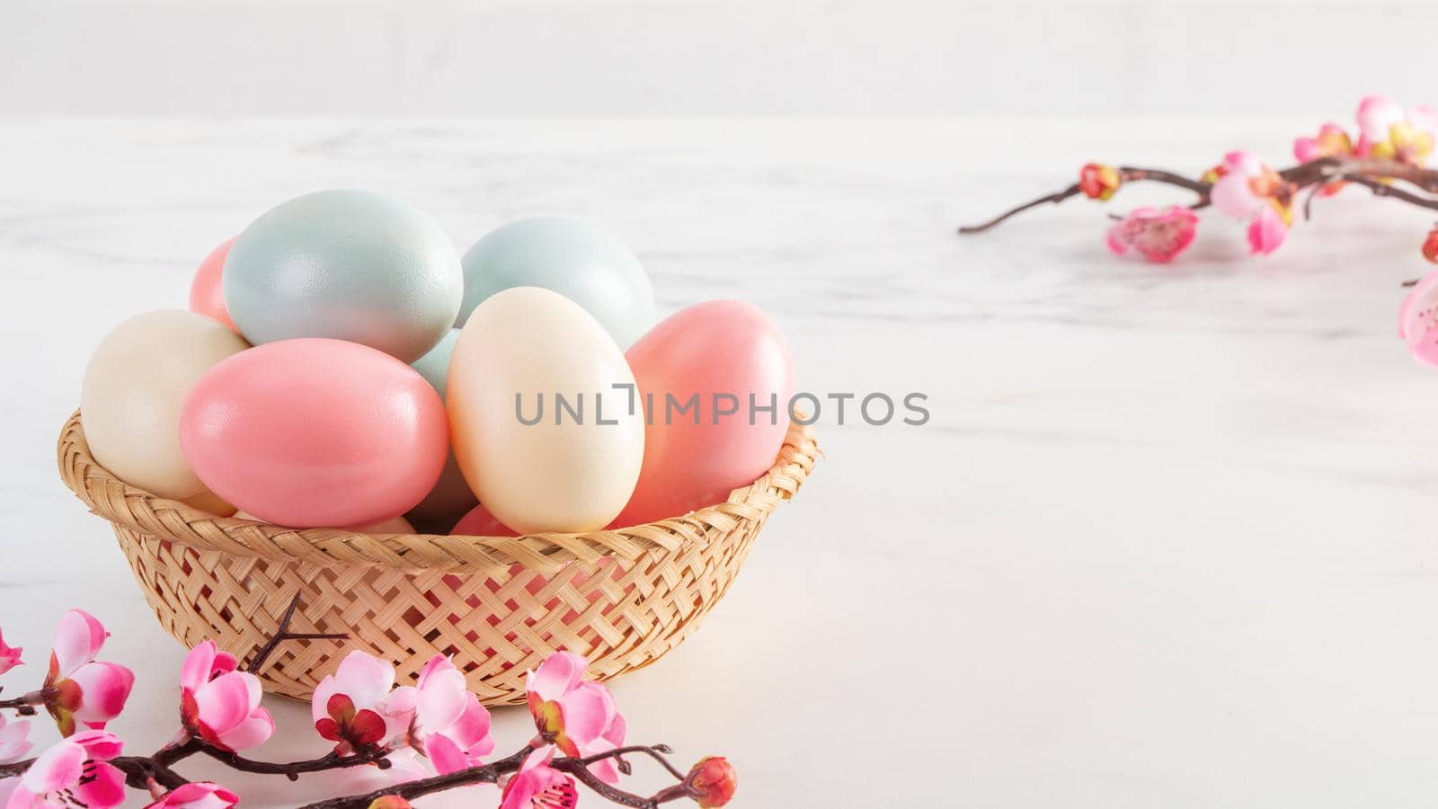 Close up of colorful Easter eggs in the basket with pink plum flower on bright white wooden table background.