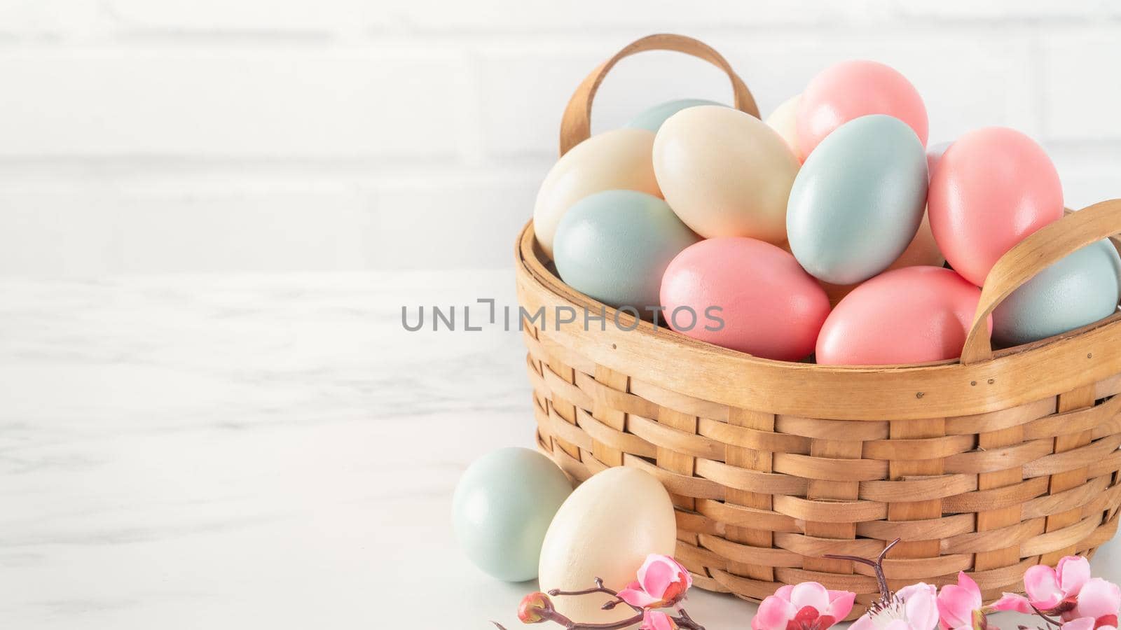 Close up of colorful Easter eggs in the basket with pink plum flower on bright white wooden table background.