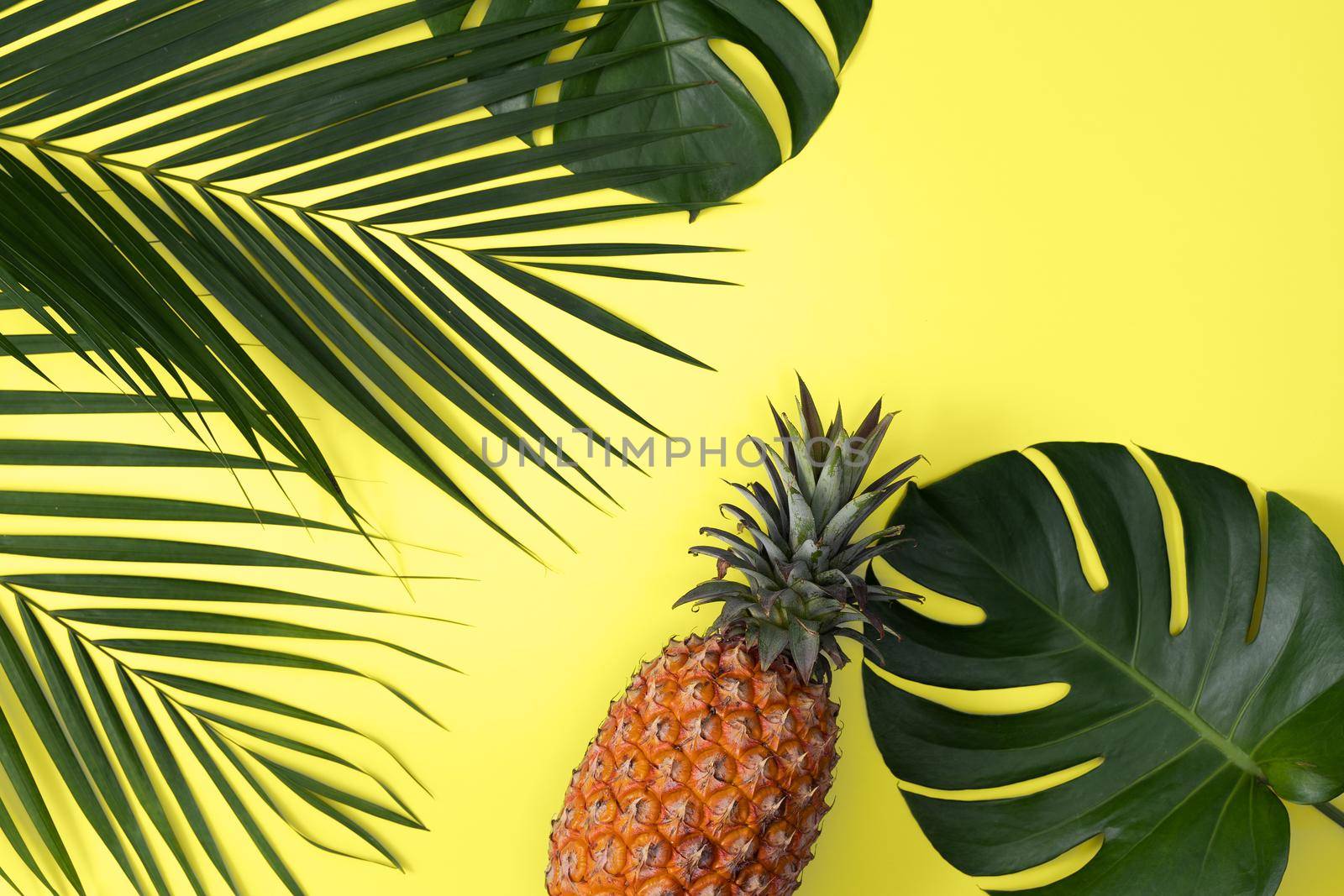 Top view of fresh pineapple with tropical palm and monstera leaves on yellow table background.