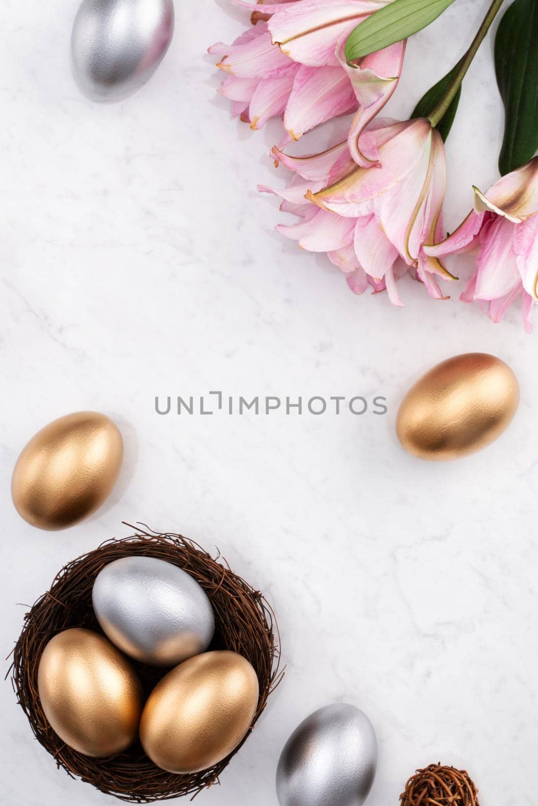 Golden and silver Easter eggs in the nest with pink lily flower on bright marble white table background.