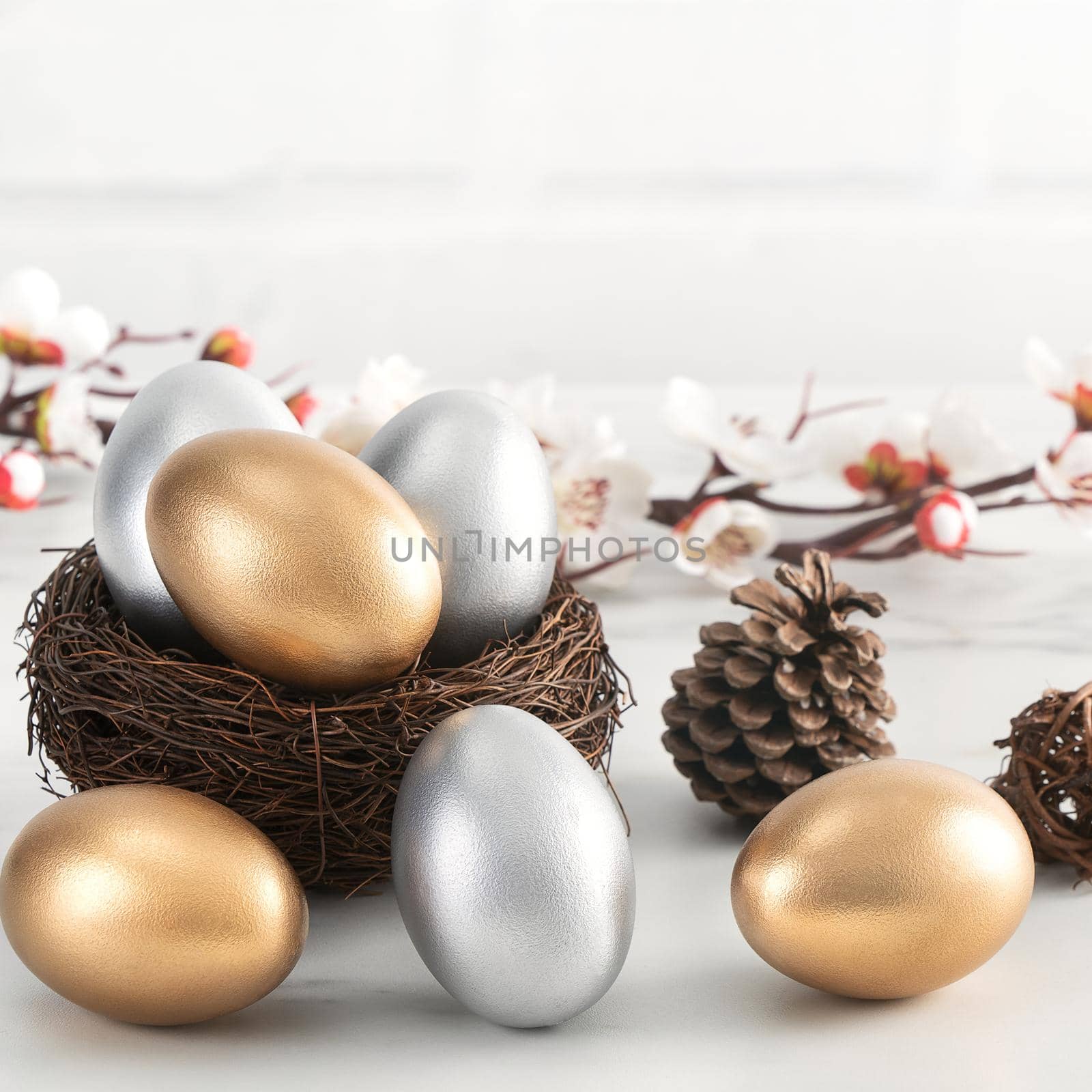 Close up of golden and silver Easter eggs in the nest with white plum flower on bright white wooden table background.