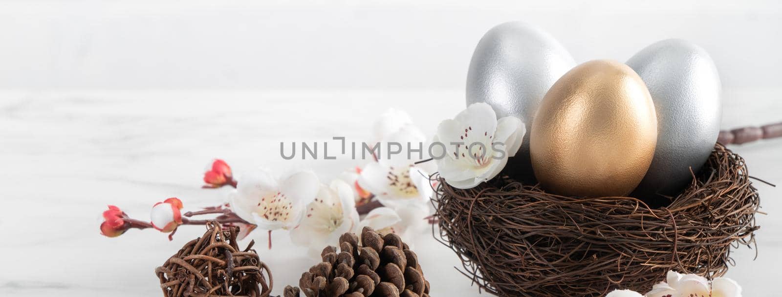 Close up of golden and silver Easter eggs in the nest with white plum flower on bright white wooden table background.