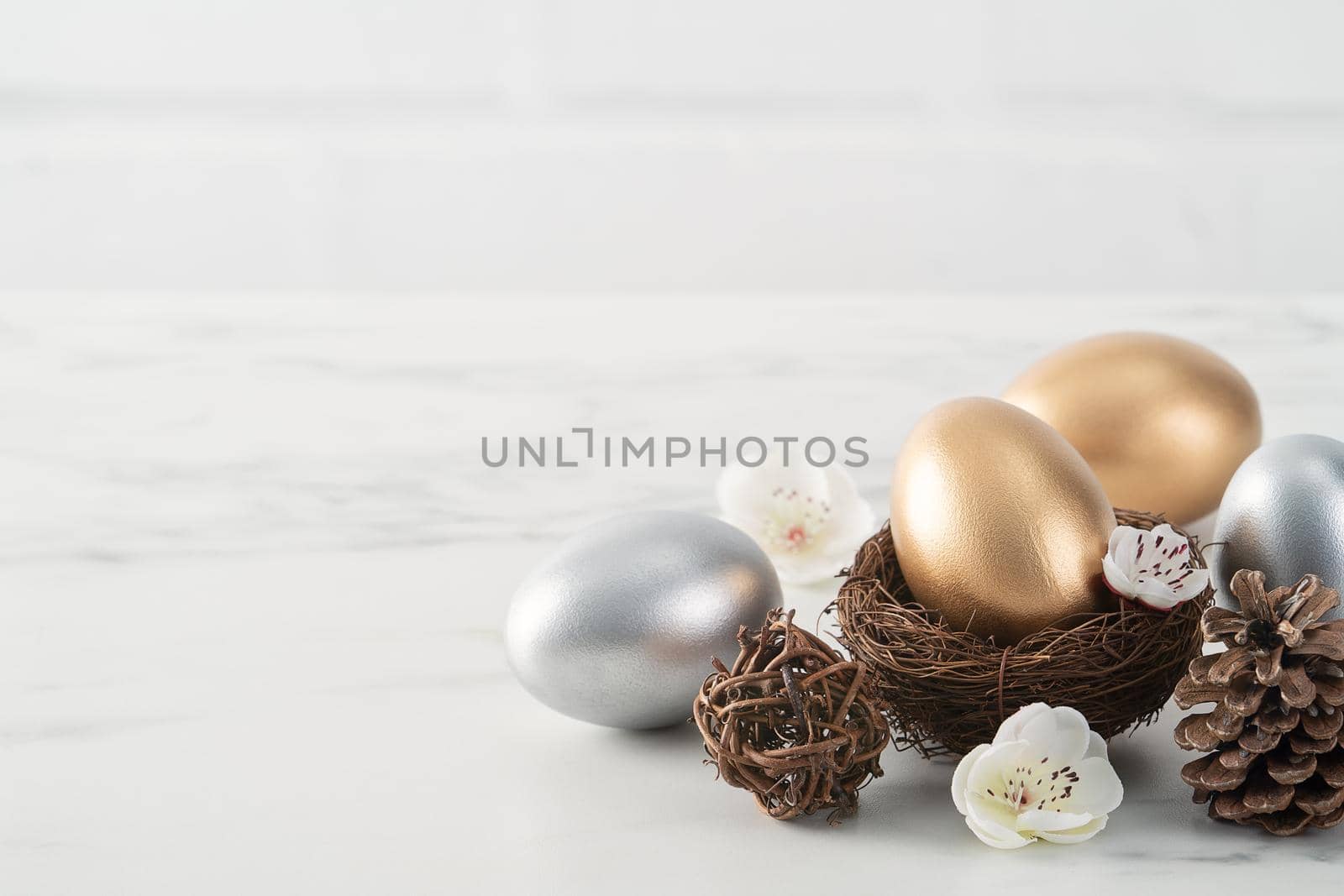 Close up of golden and silver Easter eggs in the nest with white plum flower on bright white wooden table background.