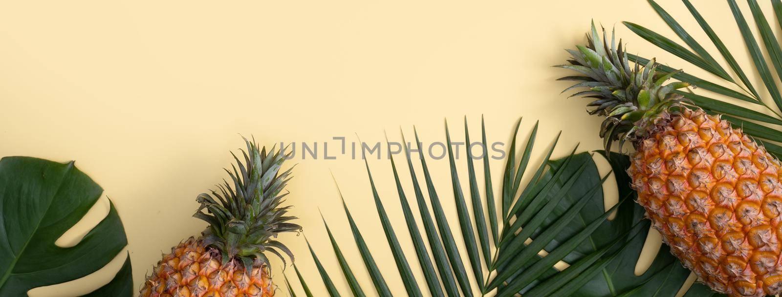 Top view of fresh pineapple with tropical palm and monstera leaves on yellow table background.