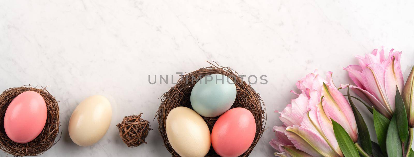 Colorful Easter eggs in the nest with pink lily flower on bright marble white table background.