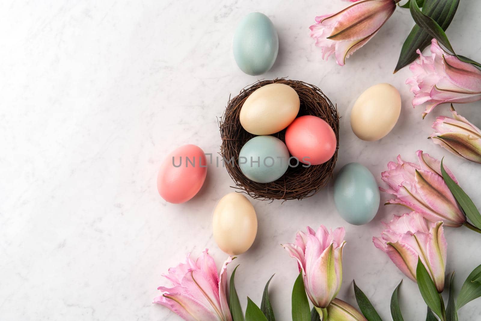 Colorful Easter eggs in the nest with pink lily flower on bright marble white table background.