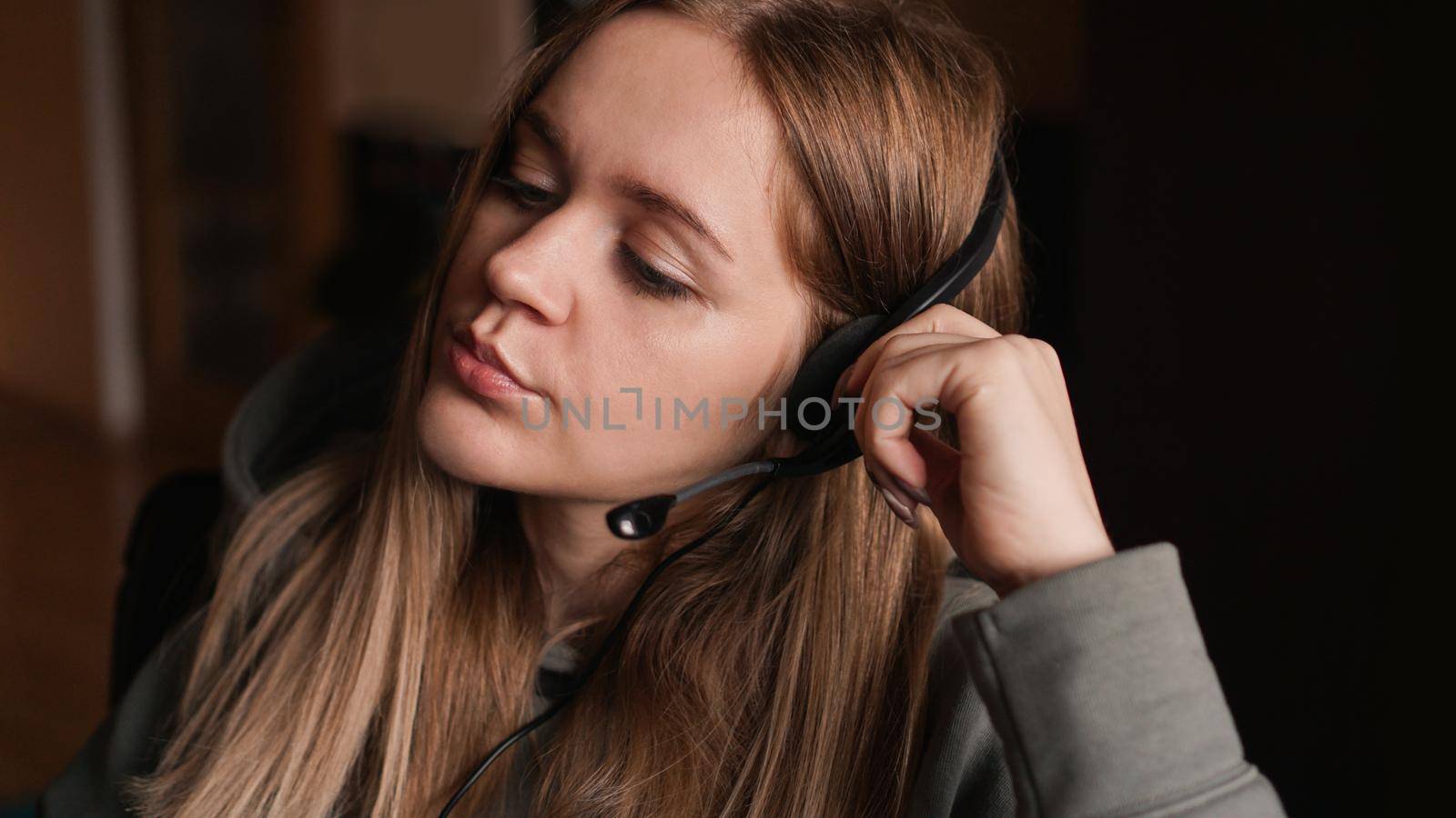 Portrait of a young girl in a hoodie and with a headset. She listens to the client with a tired look. Monotonous work. Call center worker. Remote work from home. Brown tones pictures