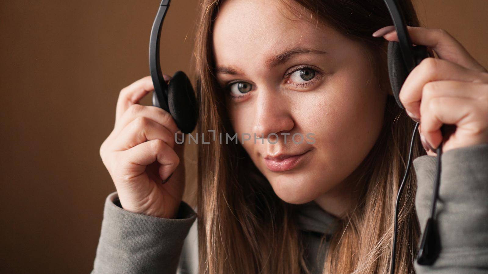 Portrait of a young girl in a hoodie puts on a headset. She looks at the camera and smiles. Call center worker. Remote work from home. Brown tones pictures