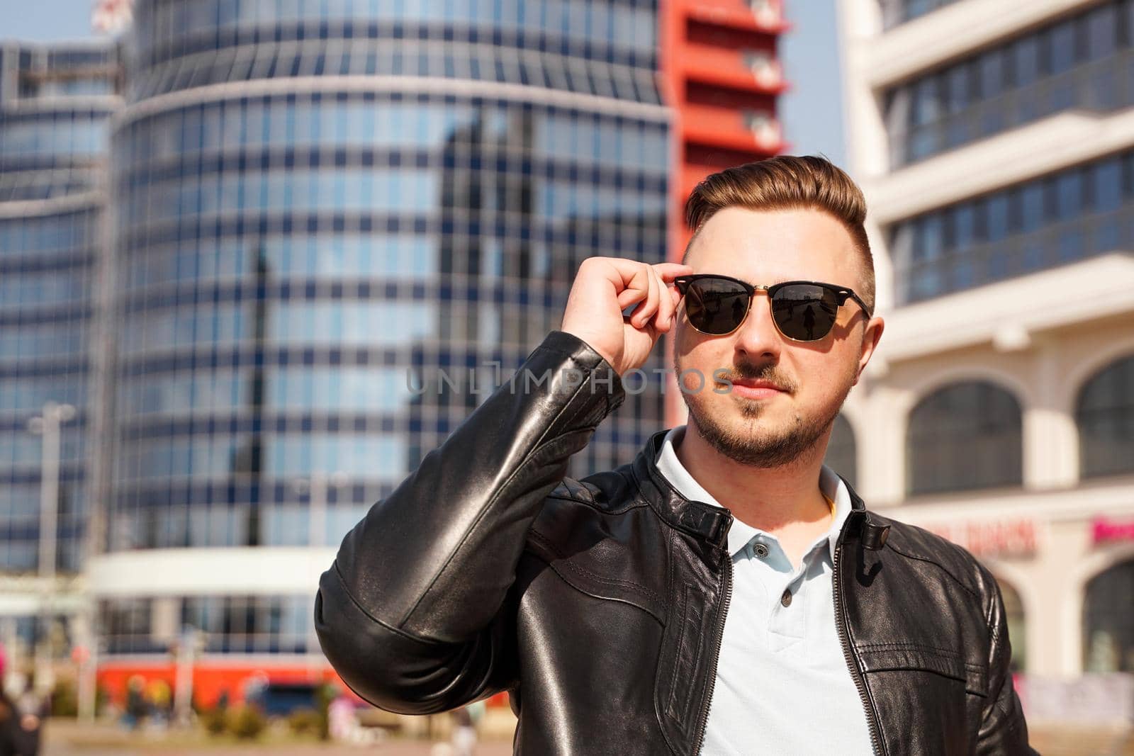 An attractive man wearing black leather jacket with dark sunglasses against the backdrop of a glass building in the city