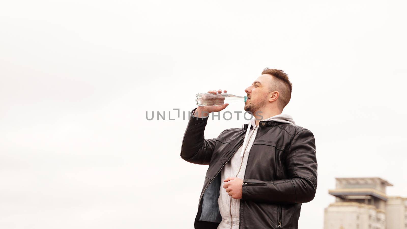 A man in a leather jacket drinks water from a bottle against the background of the sky