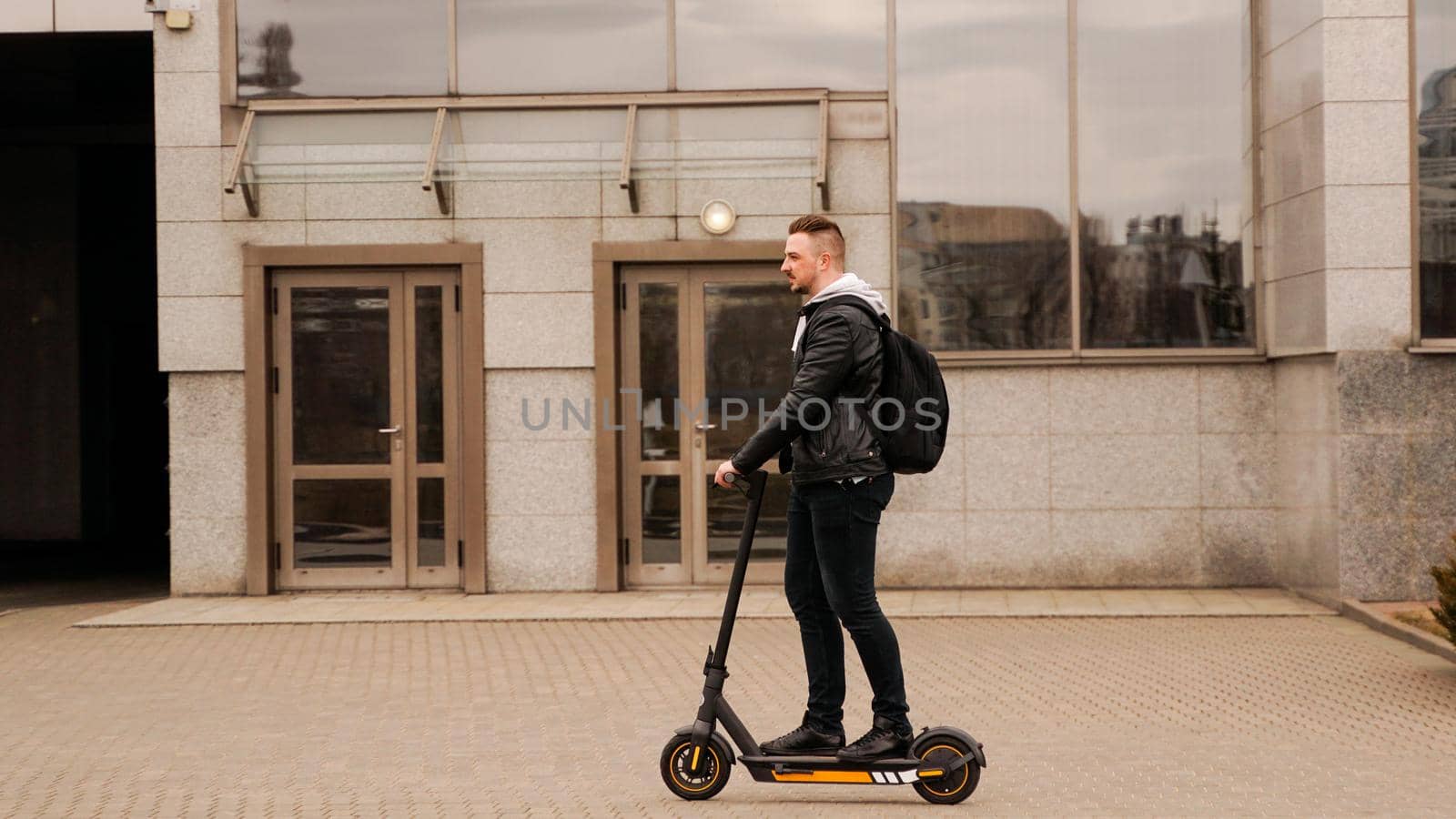 Tall man on an electric scooter against the backdrop of gray city buildings. Guy in a black leather jacket