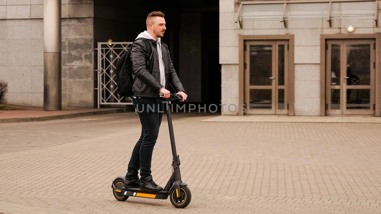 Tall man on an electric scooter against the backdrop of gray city buildings. Guy in a black leather jacket