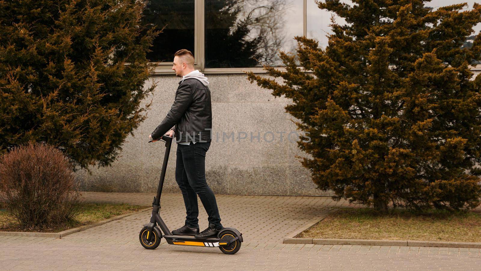 Modern man riding electric scooter in the city. A young man in a black jacket drives around the city on a sunny day.