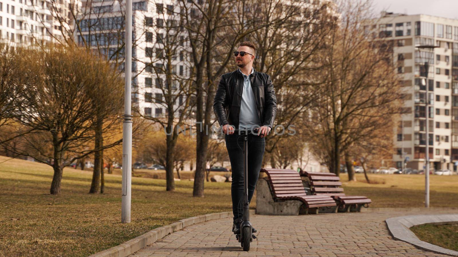 Young man in a black jacket and sunglasses rides an electronic scooter in a spring park on a sunny day