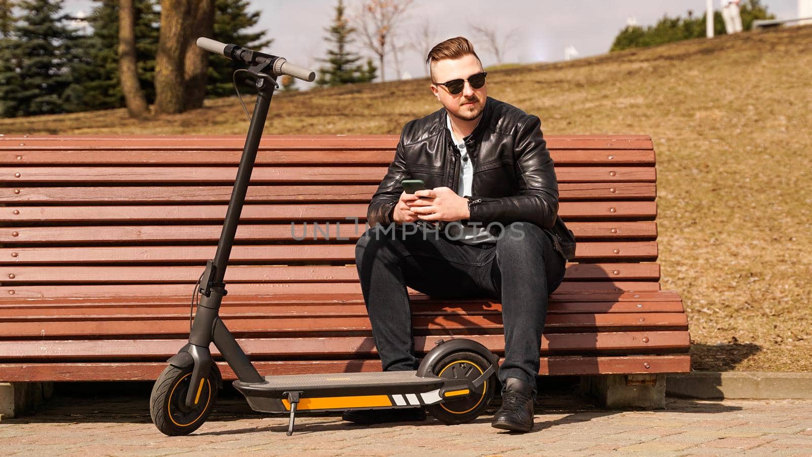 Young man sits on a bench on a spring day. Electric scooter next to a bench by natali_brill