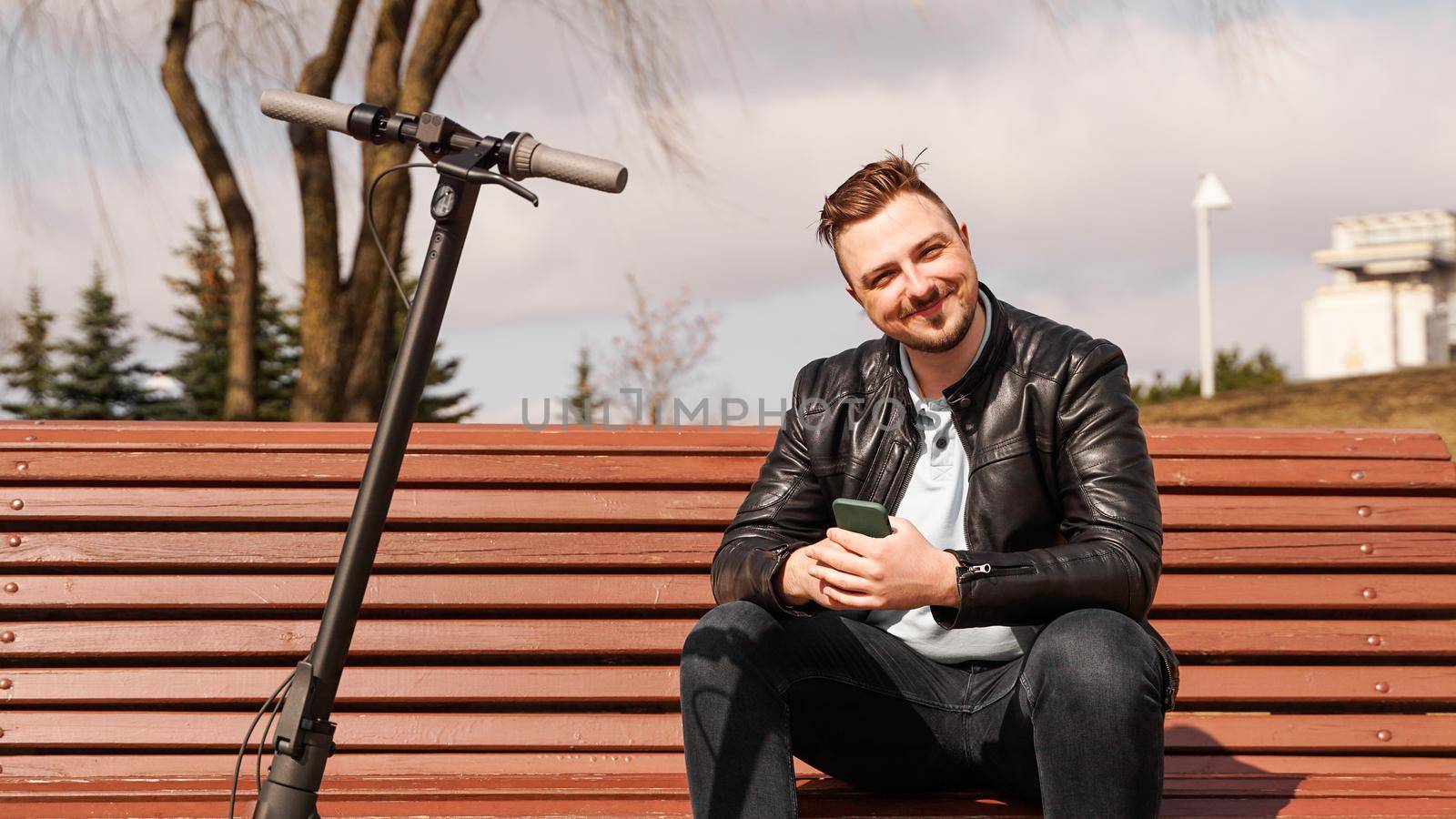 Young man sits on a bench on a spring day. Electric scooter next to a bench by natali_brill