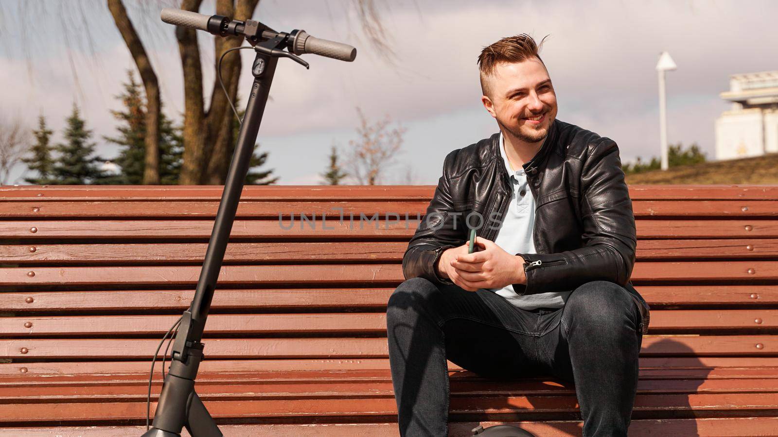 Young attractive man sits on a bench on a spring day. Electric scooter next to a bench. Man holds a smartphone in his hands. He looks away and smiles