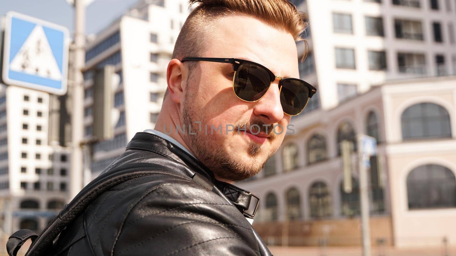 An attractive man wearing black leather jacket with dark sunglasses against the backdrop of a glass building in the city