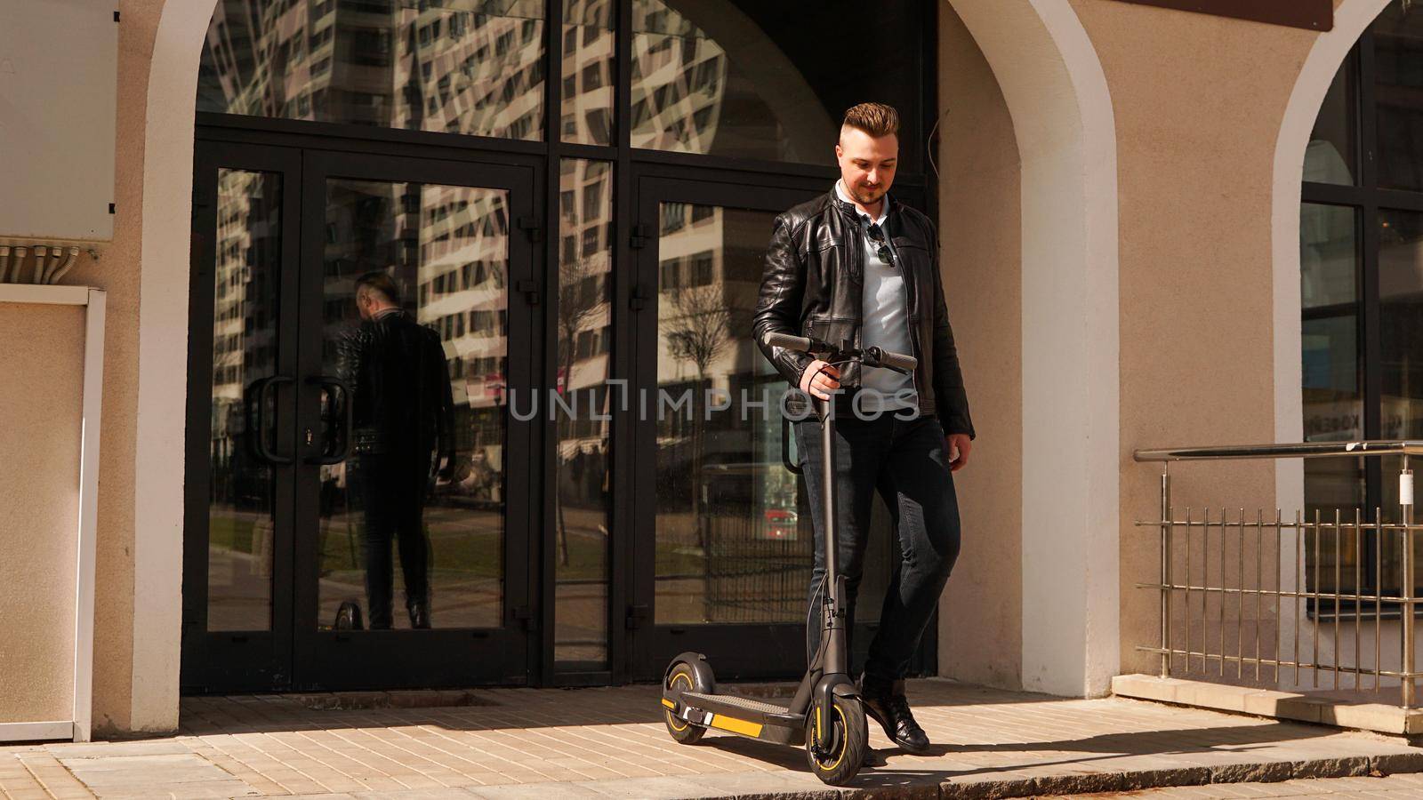 A young man comes out of the entrance of his house and holds an electric scooter in his hand. He is going to ride a scooter. Sunny day.