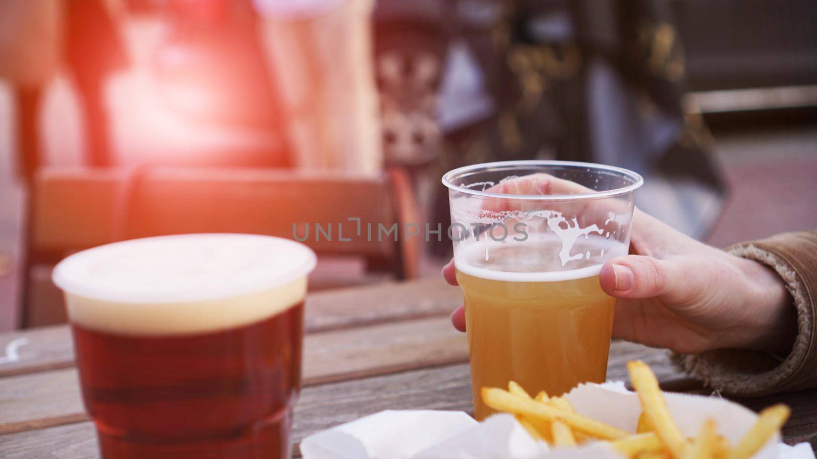 Girlfriends drink beer at the street food festival. Plastic glass of beer in hand. French fries, unhealthy food. Glare of the sun